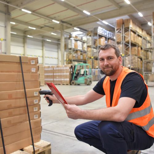 A man wearing fluorescent orange high visibility clothing scanning barcodes on cardboard boxes in a warehouse