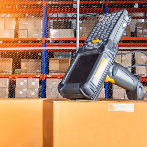 Hand held silver barcode scanner machine, sat on a cardboard box in front of warehouse racking