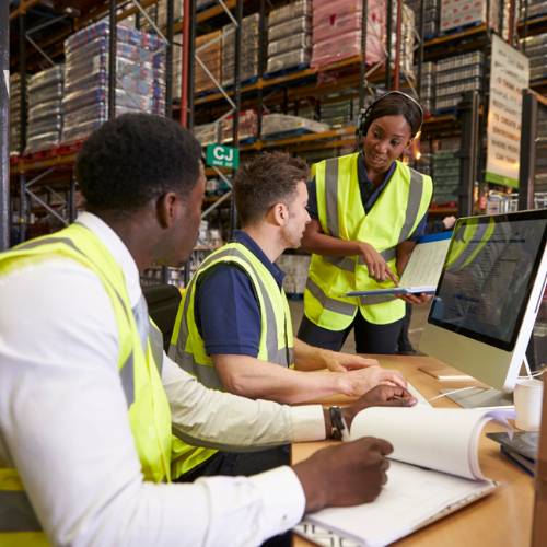 Three people in a warehouse working on a computer and checking items off items on a pad of paper