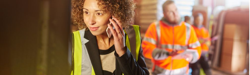 A woman on the phone wearing a yellow high vis jacket, men stand behind her in orange high vis jackets. They are surrounded by cardboard boxes