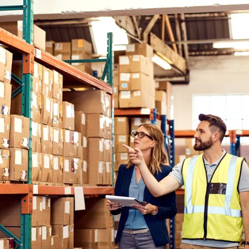 People in a warehouse pointing at cardboard boxes stocked on industrial racking
