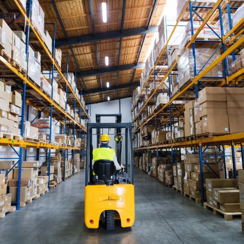A person wearing yellow high vis personal protective clothing driving a forklift truck through a warehouse with industrial racking storing stock