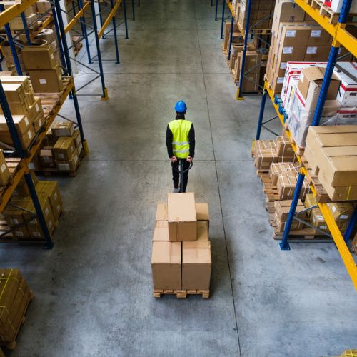 Someone in a warehouse wearing personal protective equipment clothing pulling along a pallet truck containing cardboard boxes