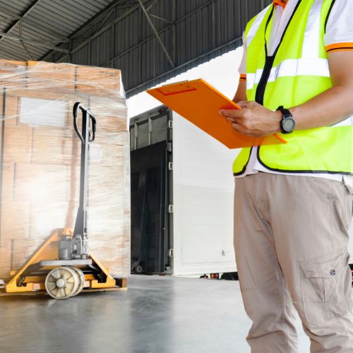Warehouse worker checking stock on a clipboard, with pallet truck in background piled high with cardboard boxes