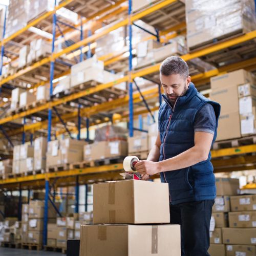 Warehouse worker taping cardboard boxes up, with industrial shelving in the background filled with packaged stock