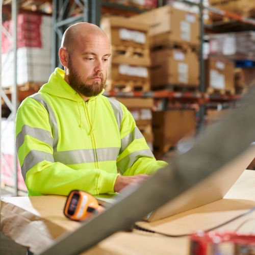 A member of staff in a warehouse working on a laptop with a handheld scanner on the desk