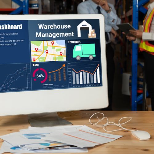 Computer on a desk with a screen saying Warehouse Management, two workers in the background holding a computer tablet