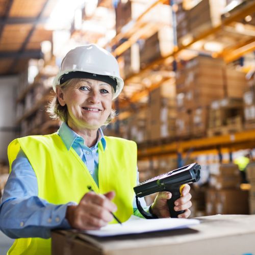 A warehouse worker in yellow high visibility clothing and hard hat holding a handheld scanner ticking off items on a sheet of paper