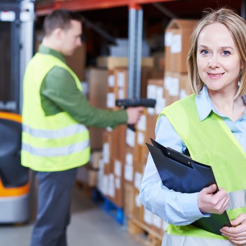 Two warehouse workers in yellow high visibility clothing, one scanning boxes, one looking at the camera holding a clipboard