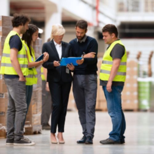 Group of people talking with a woman in a warehouse, some are wearing high vis jackets, they're discussing business documents
