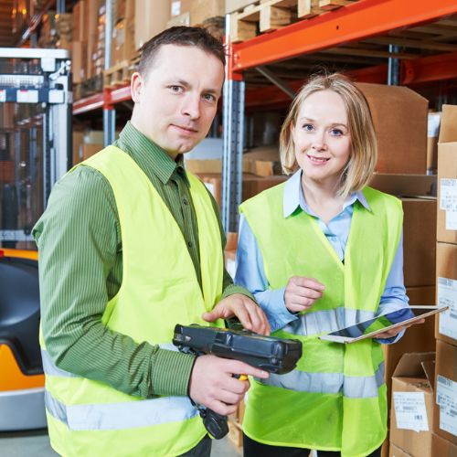 Two warehouse workers in yellow high visibility clothing, one scanning boxes, one looking at the camera holding a clipboard