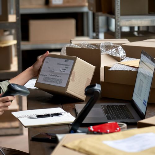 A warehouse worker sitting at a desk scans a barcode on a cardboard box with a laptop in front of them
