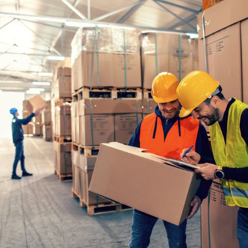 Warehouse workers in high visibility clothing and hard hats holding cardboard boxes and checking off items on a list