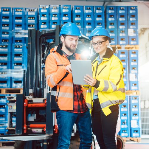 Warehouse workers in high visibility clothing and hard hats checking items on a computer tablet, in front of blue crates and a forklift truck