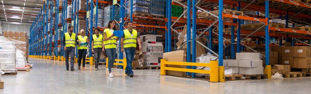 A group of people walk through a warehouse stacked with stock, they wear high vis jackets