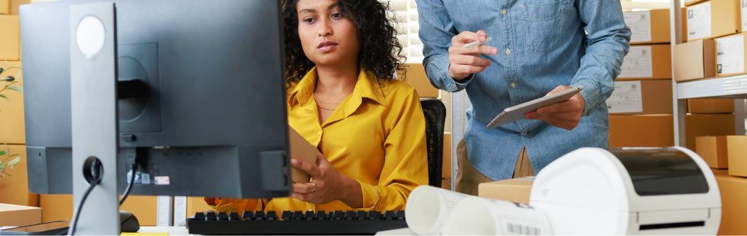 A person sits at a computer screen, with a lable printer in front and a person holding a digital tablet standing behind